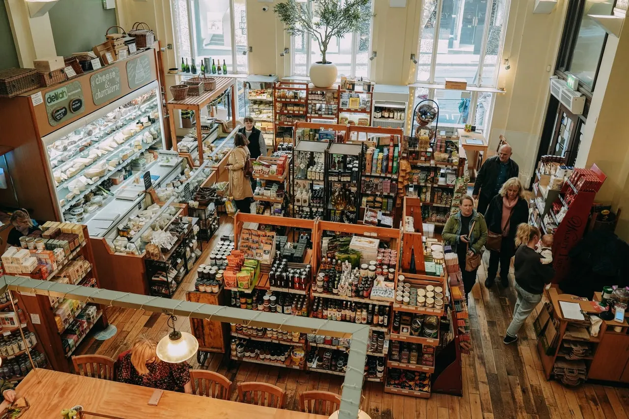 Overhead view of a delicatessen shop with customers browsing wooden display counters stocked with packaged foods, jars and provisions.
