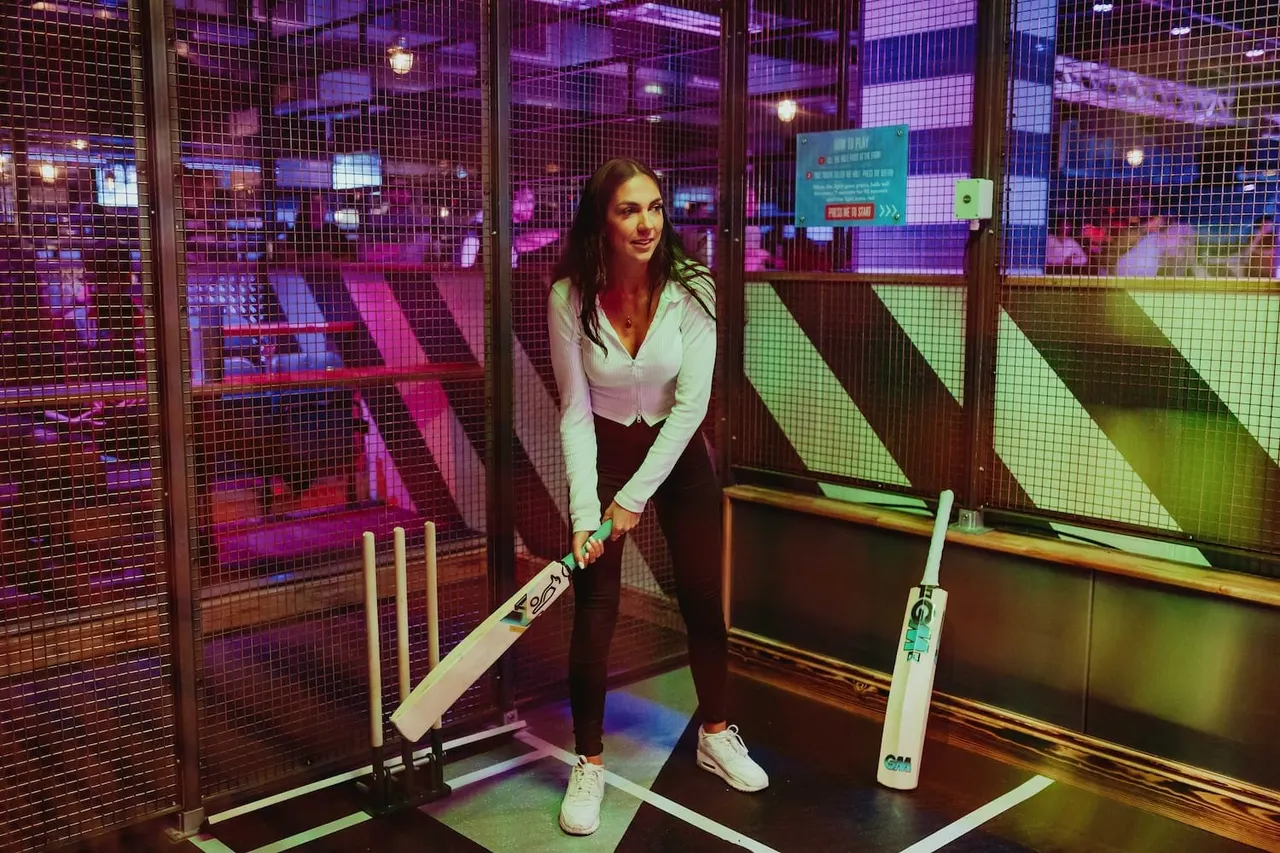 Woman holding a cricket bat in a neon-lit indoor cricket bay with purple and green lighting.
