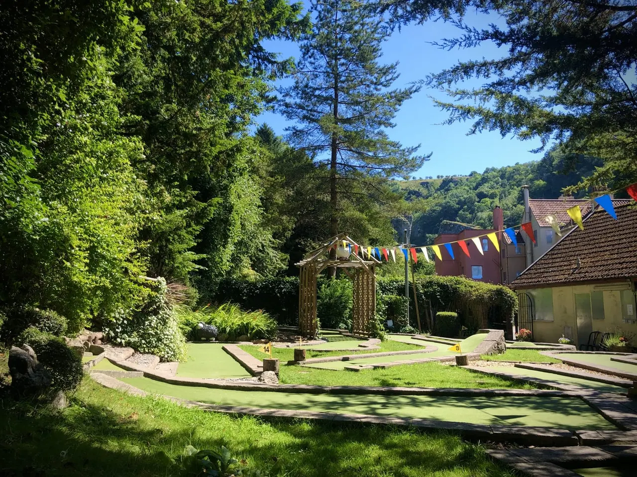 Mini golf course with bright green playing surface, wooden arbour, and colourful bunting, surrounded by trees and buildings.
