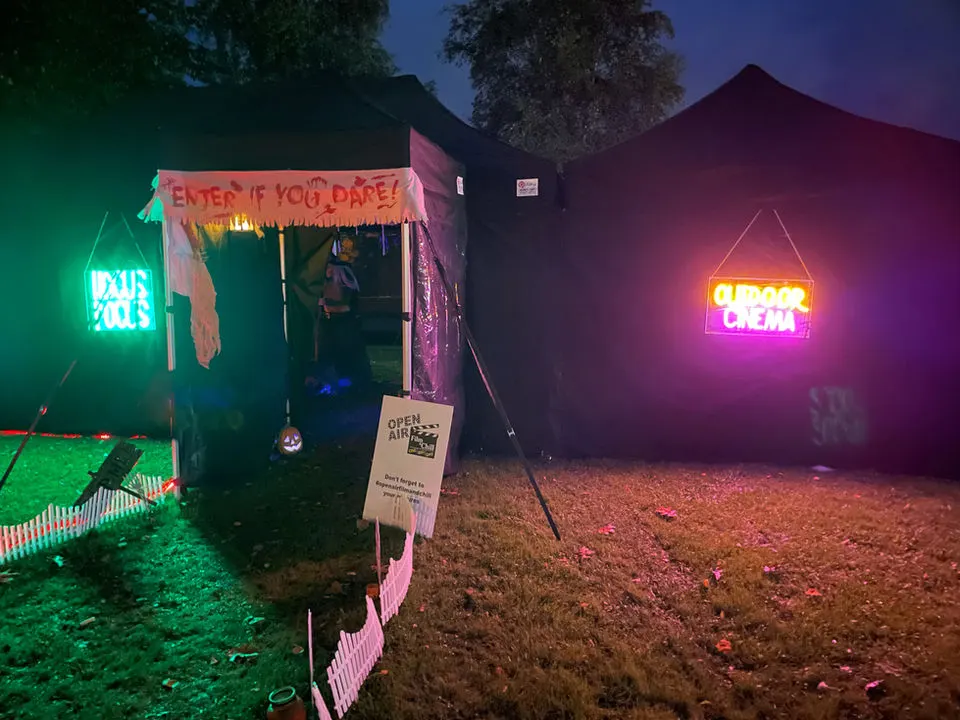 Outdoor cinema venue at dusk with illuminated neon signs on two dark tents, featuring "Enter if you dare" banner and pink fencing around the grounds.