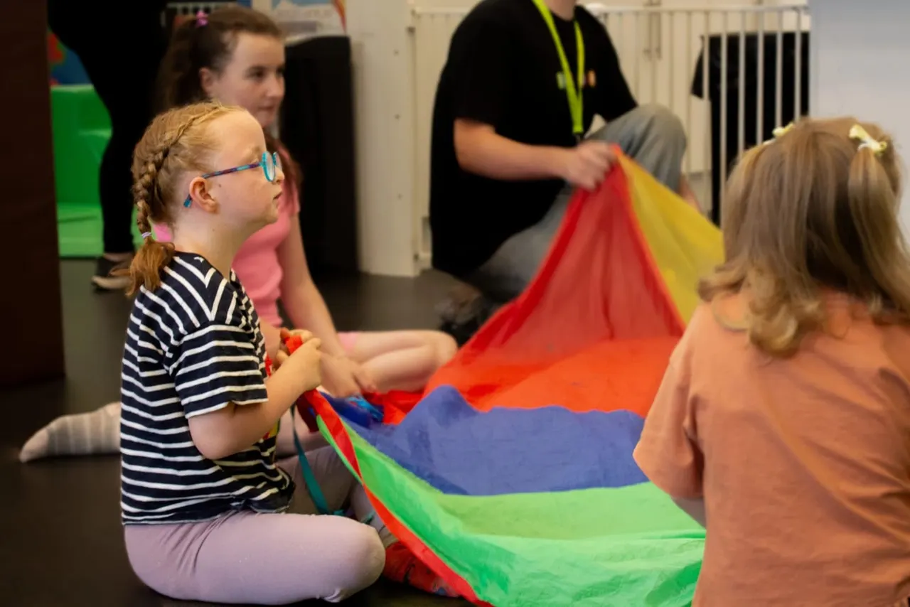 Children holding a colourful parachute during an indoor activity session with staff supervising.