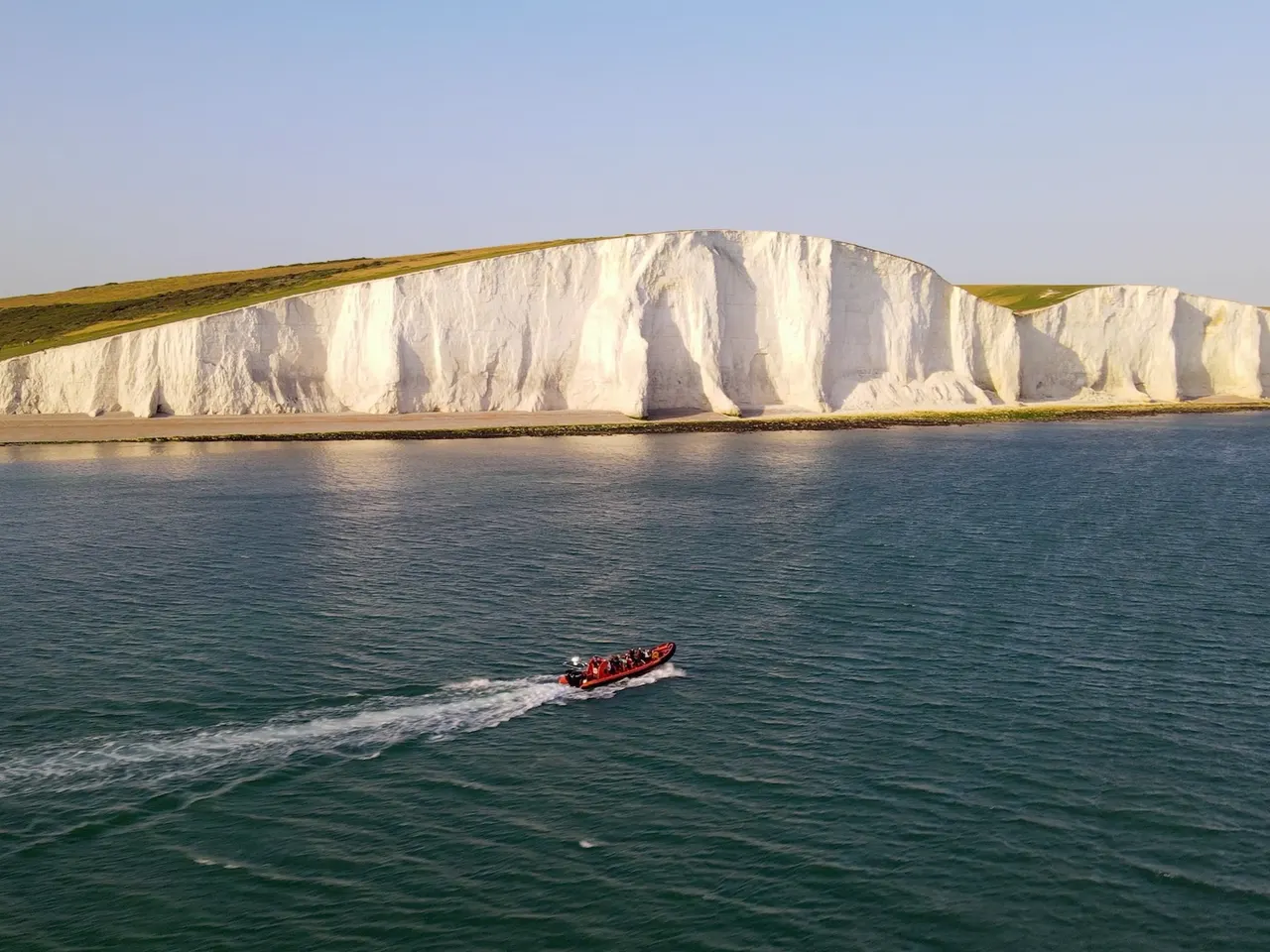 Speedboat full of passengers approaching tall white chalk cliffs across turquoise sea water.