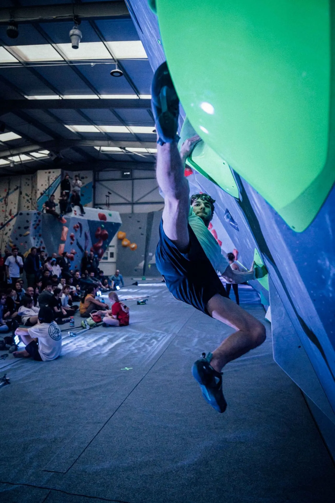 A climber ascending an indoor climbing wall with spectators watching from below in an indoor facility.