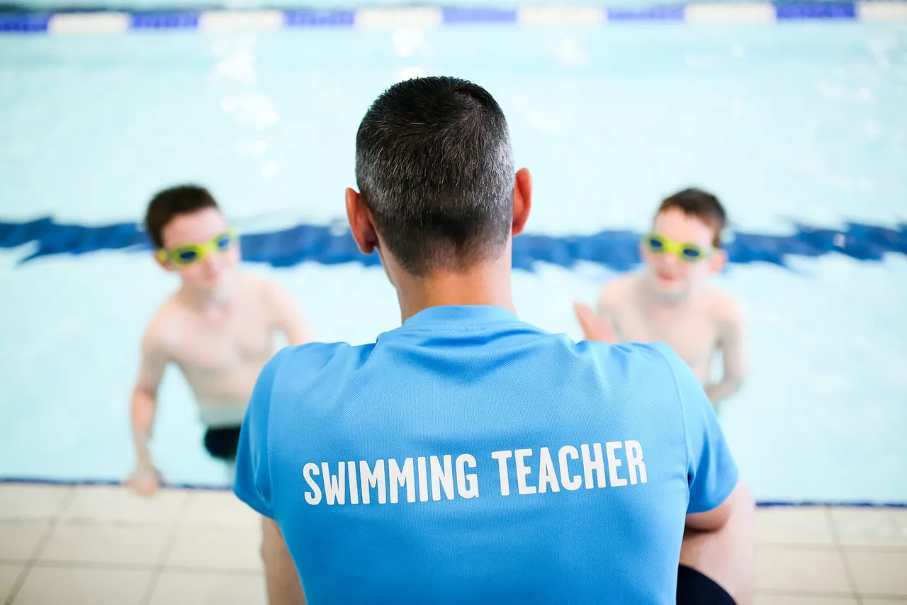 Swimming teacher in blue shirt instructs two young children wearing yellow goggles in an indoor pool.