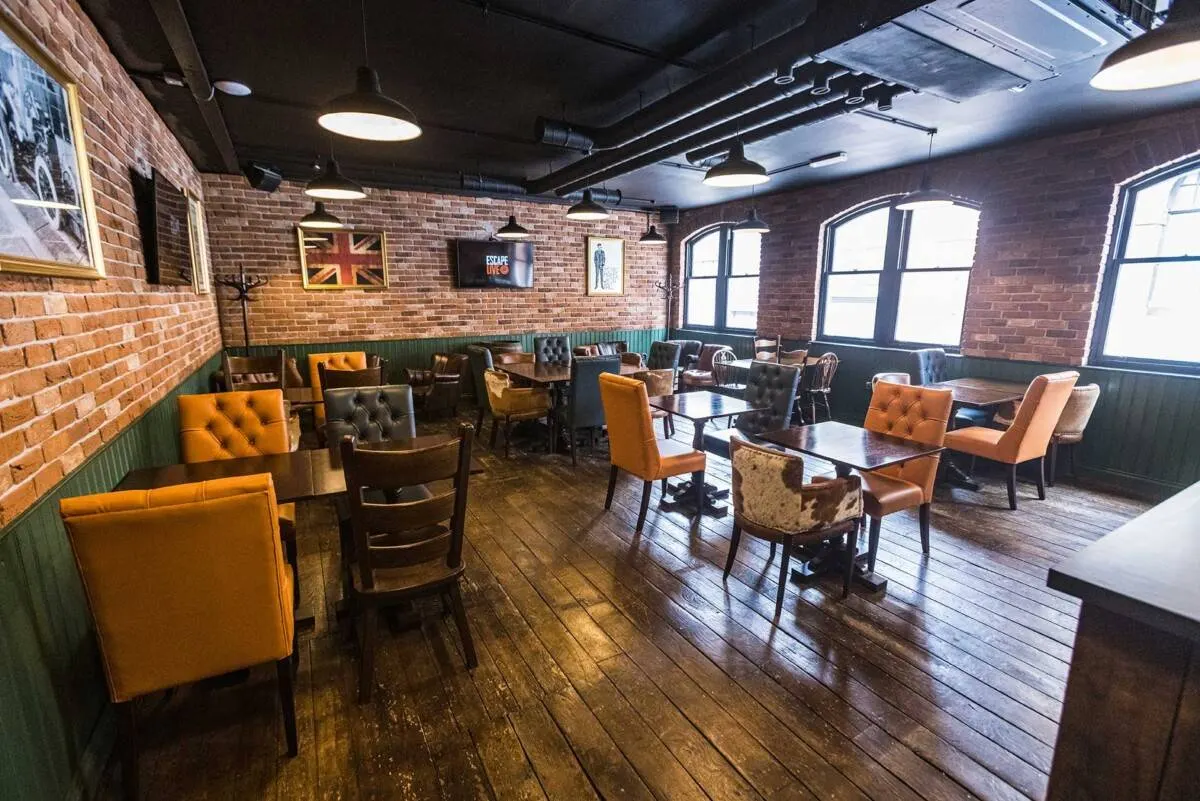 Interior of an escape room venue with brick walls, orange and black chairs at tables, and industrial-style lighting.