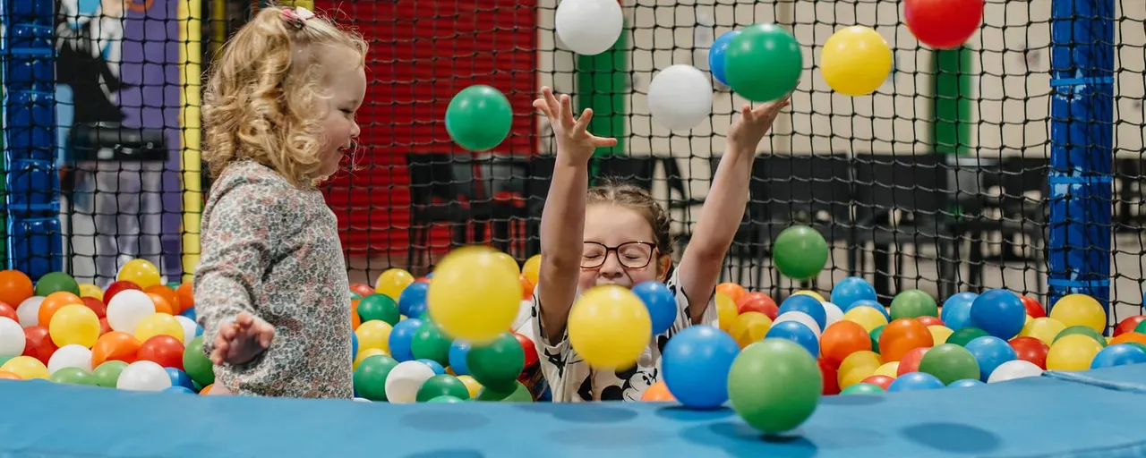 Two young children playing in a colourful ball pit with suspended balls above them.