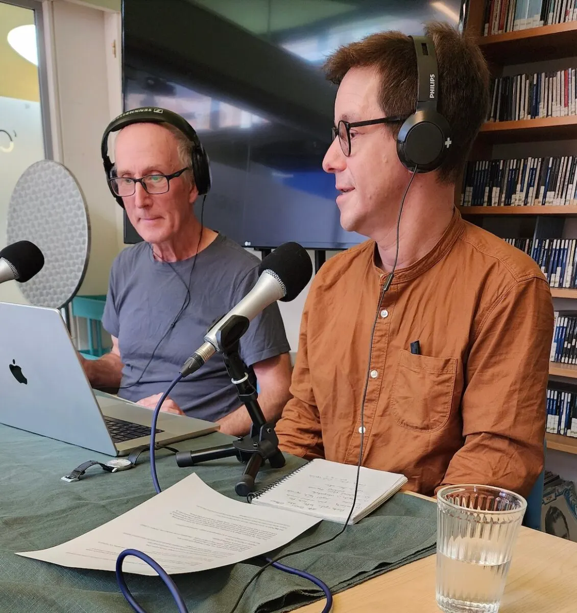 Two men recording a podcast or audio programme at a table with microphones and headphones in a library setting.