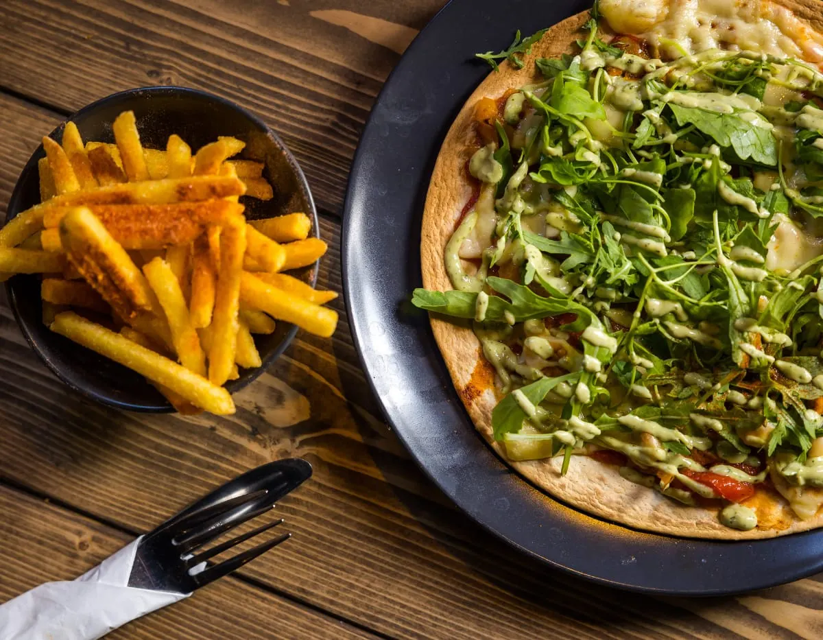 A sandwich topped with green leaves and sauce served on a dark plate alongside chips in a bowl on a wooden table.