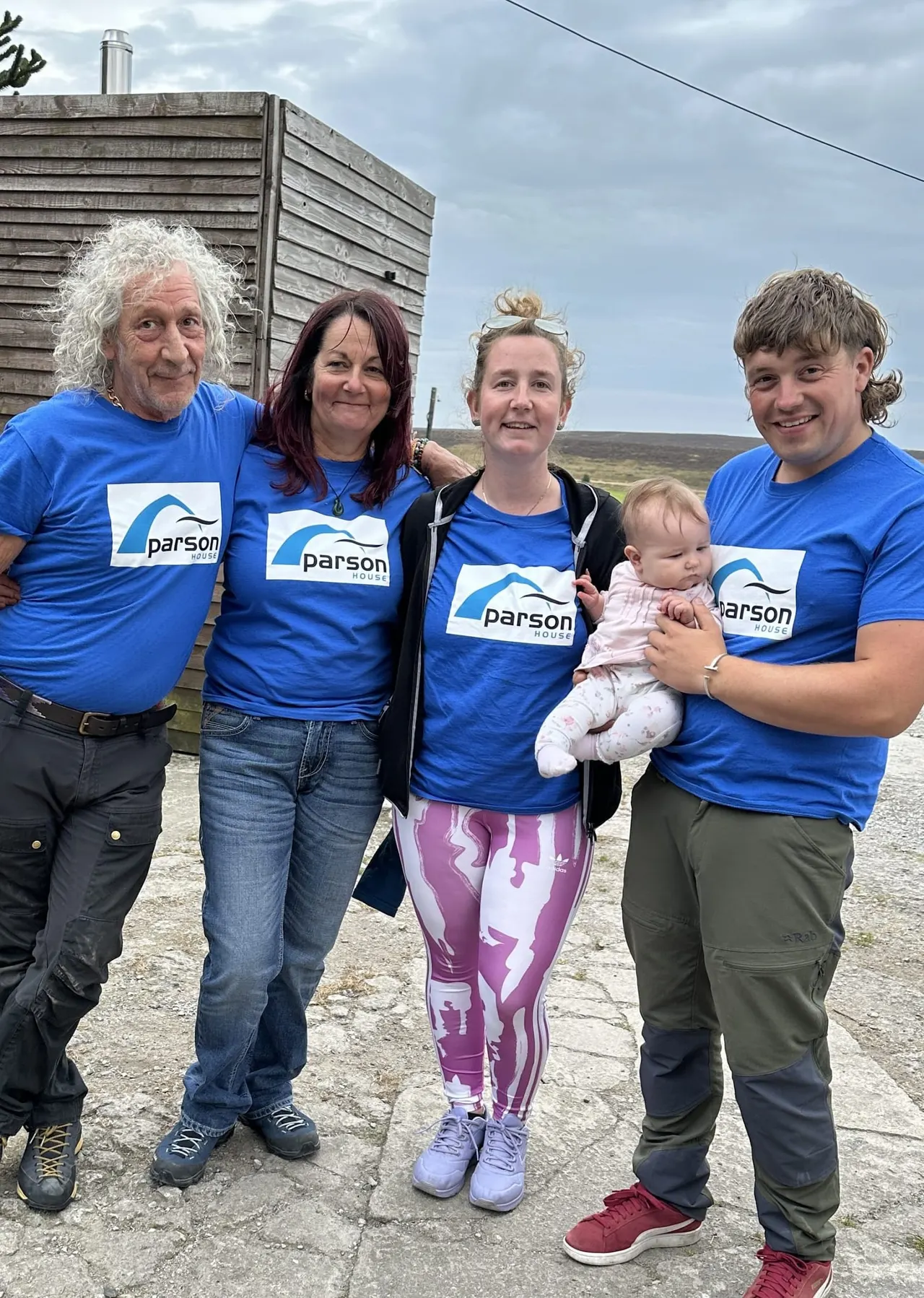 Five people wearing matching blue Parson House t-shirts stand together outdoors with moorland in the background, one holding a baby.