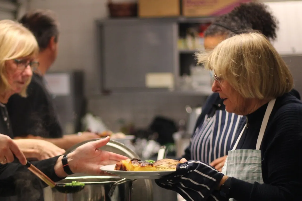 A woman in a striped apron serves food on a plate to a guest at an indoor community event.