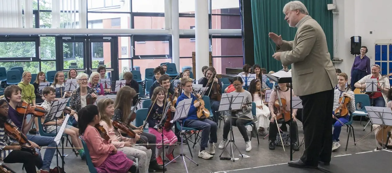 An older man conducts a large youth orchestra in a school hall, with young musicians holding violins and other string instruments seated around him.