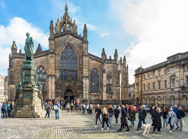 Historic stone cathedral with ornate spires and large windows, visitors gathered in the cobbled square beside a green statue.