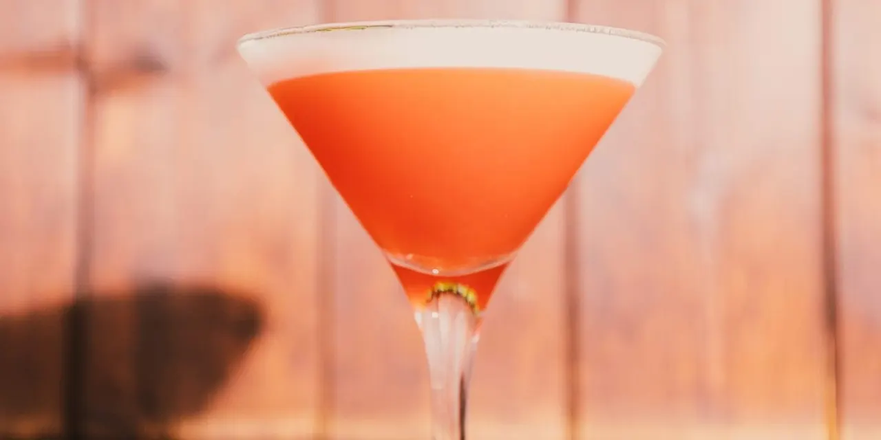 A bright orange-red cocktail in a martini glass with a sugared rim against a blurred peachy background.