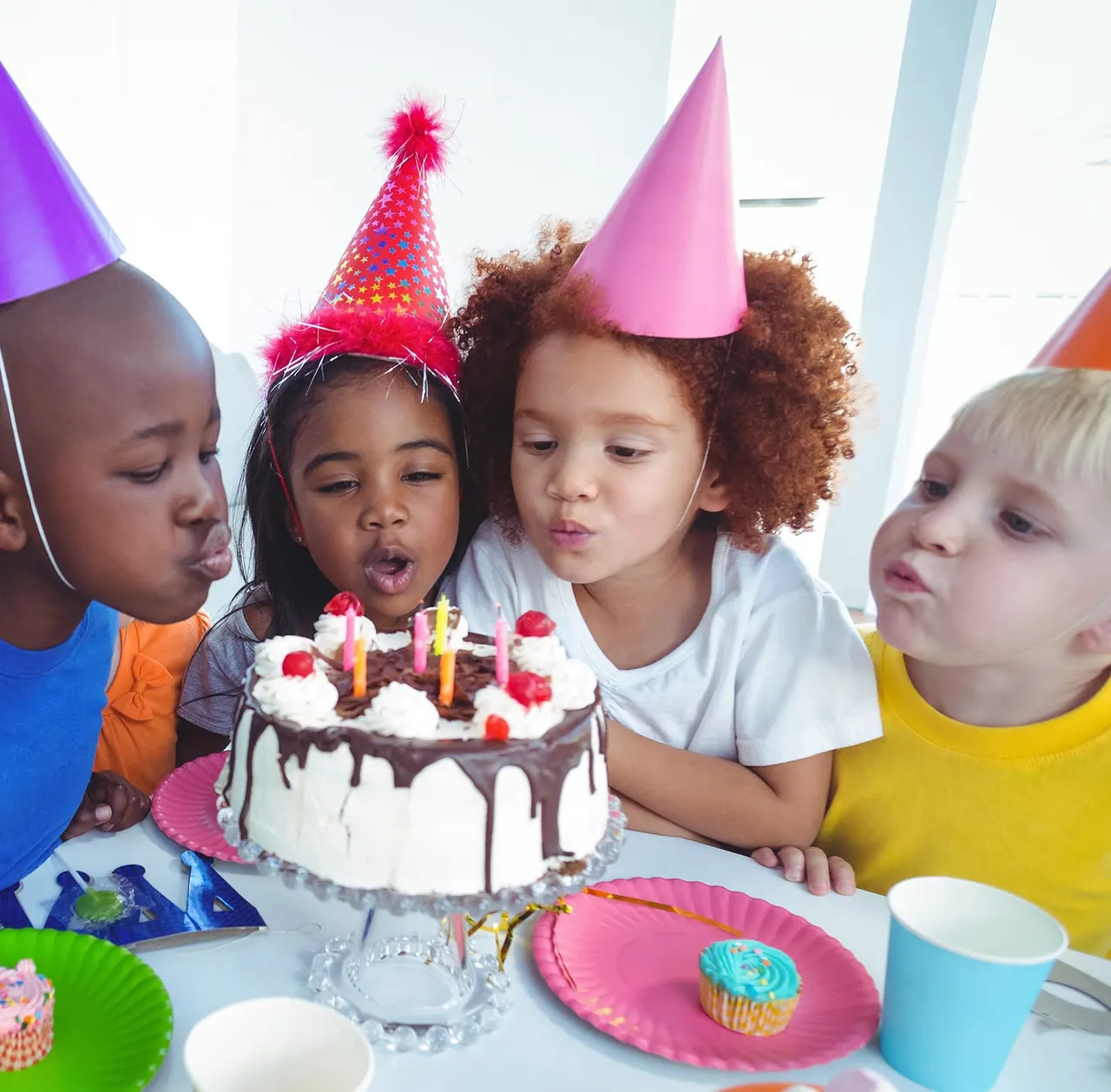 Four children wearing colourful party hats blowing out candles on a chocolate birthday cake.
