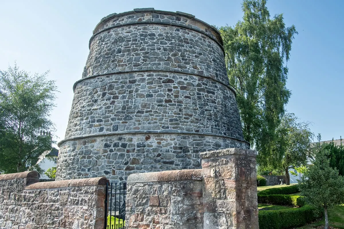 Historic cylindrical stone dovecot tower with metal bands, surrounded by trees and grounds on a clear day.