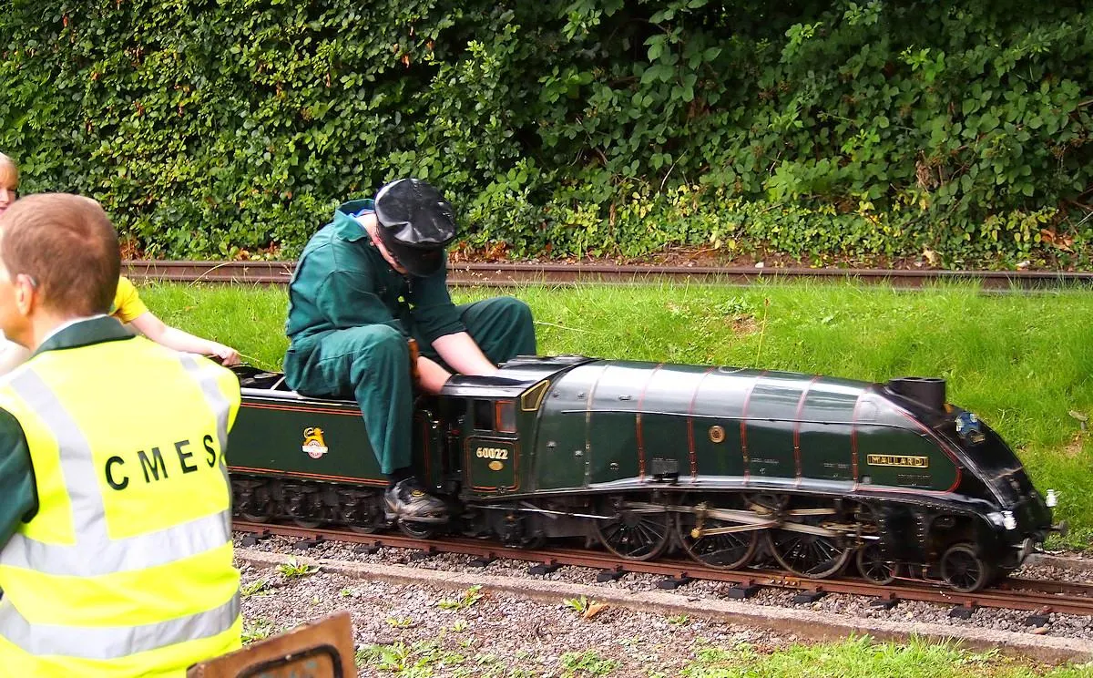 Staff members operate a miniature steam locomotive on a narrow-gauge railway track with ivy-covered fencing in the background.