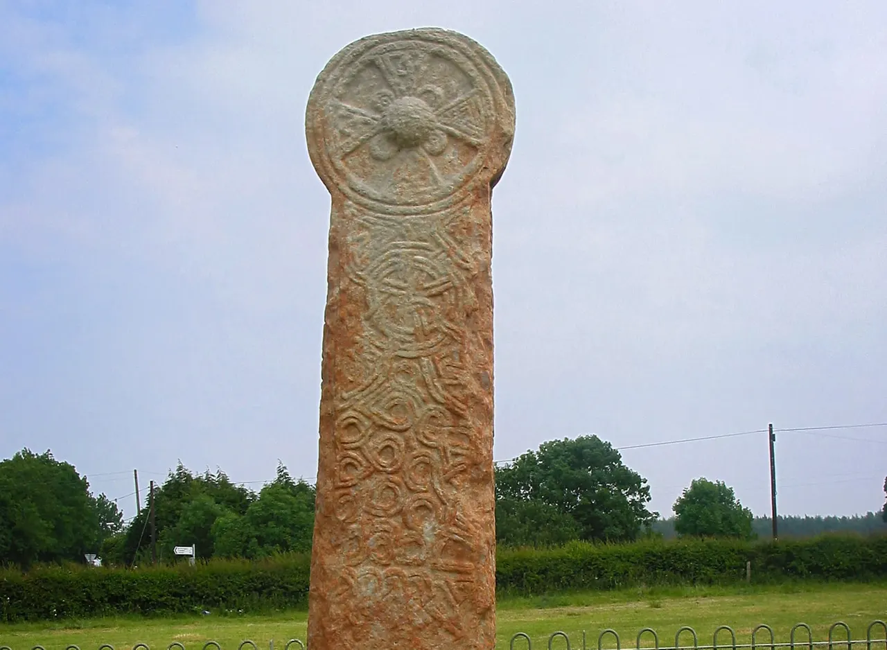 Ancient carved stone pillar with Celtic designs and circular head, displayed outdoors with trees and countryside in background.