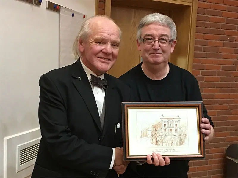 Two men shaking hands while holding a framed architectural drawing indoors.