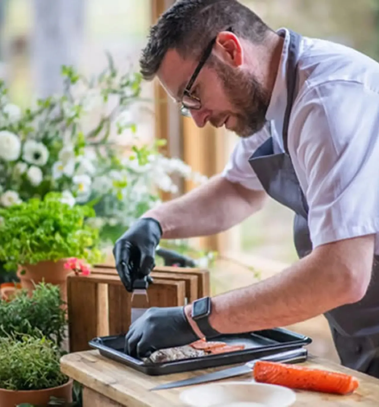 Man in apron and gloves preparing food outdoors at a table surrounded by potted plants and flowers.
