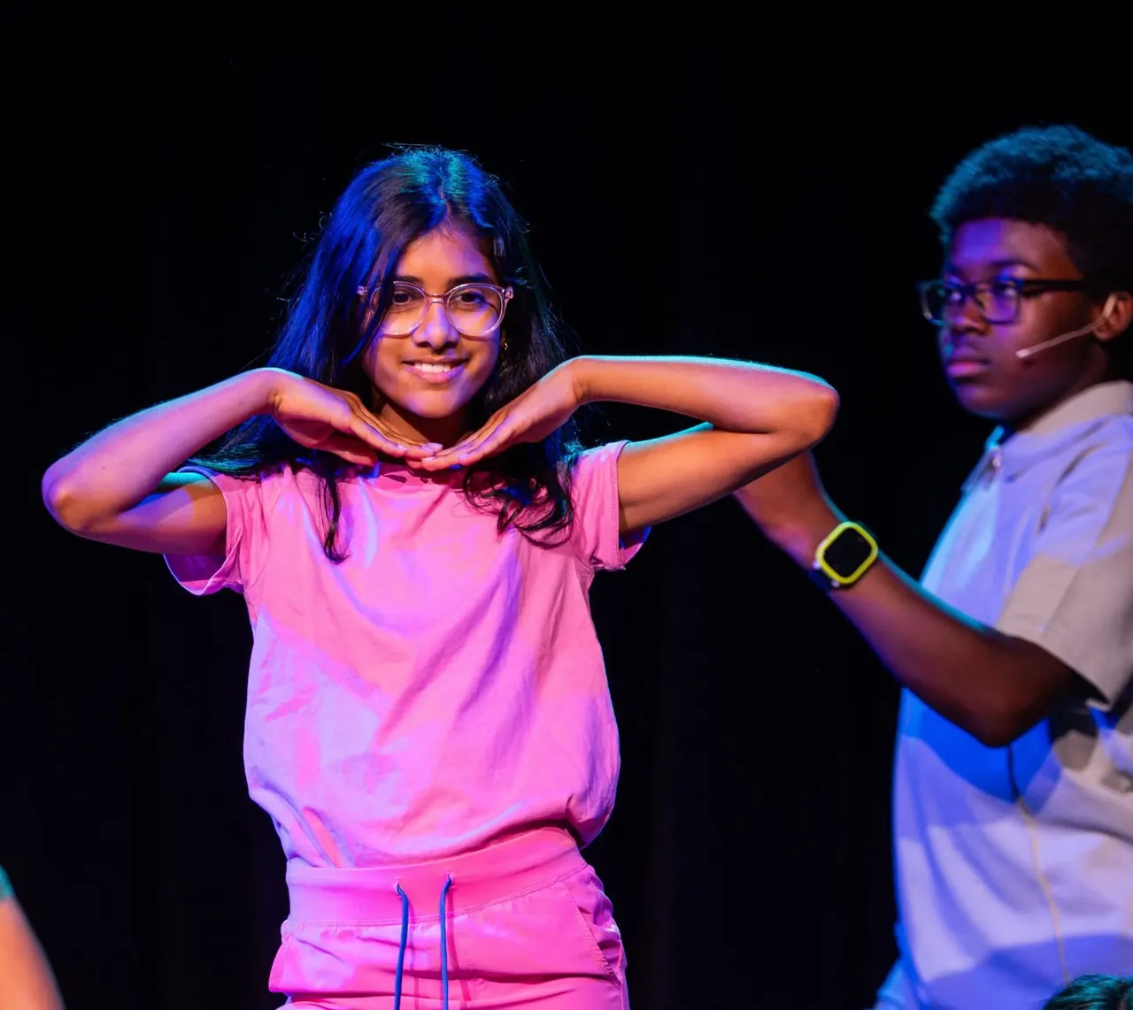 Two young performers on stage under coloured lighting, one in a pink outfit making a heart shape with their hands, smiling at the audience.
