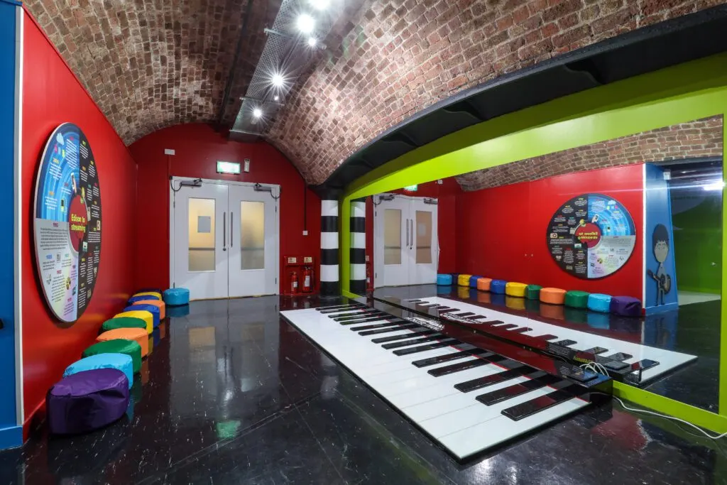 Interactive music room with a large floor piano and colourful bean bags beneath a brick arched ceiling.
