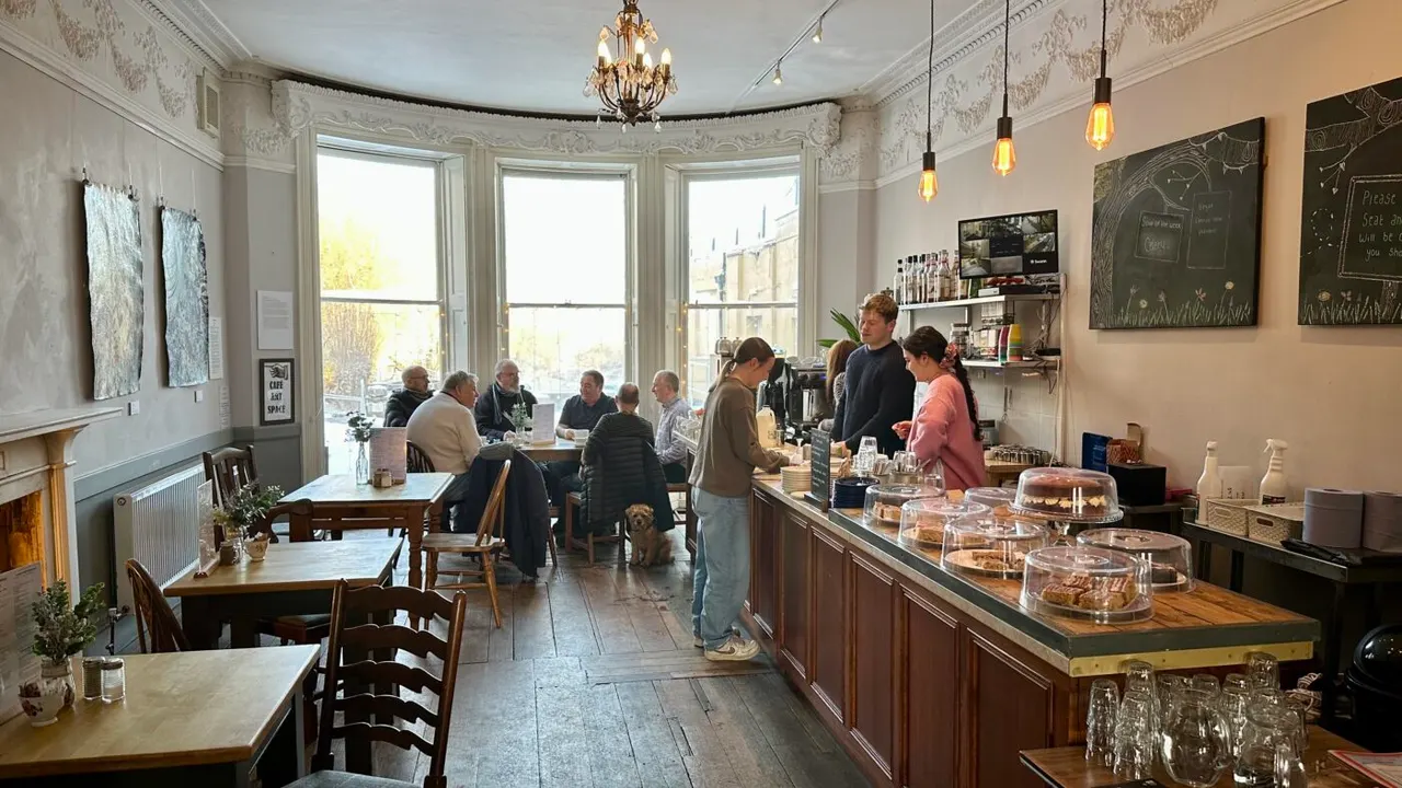 A café interior with elegant period features, customers seated at tables by bay windows, and staff behind a counter with cakes and refreshments displayed.
