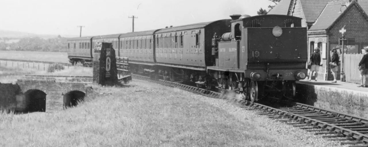 Steam locomotive with passenger carriages at a rural railway station with passengers on the platform.