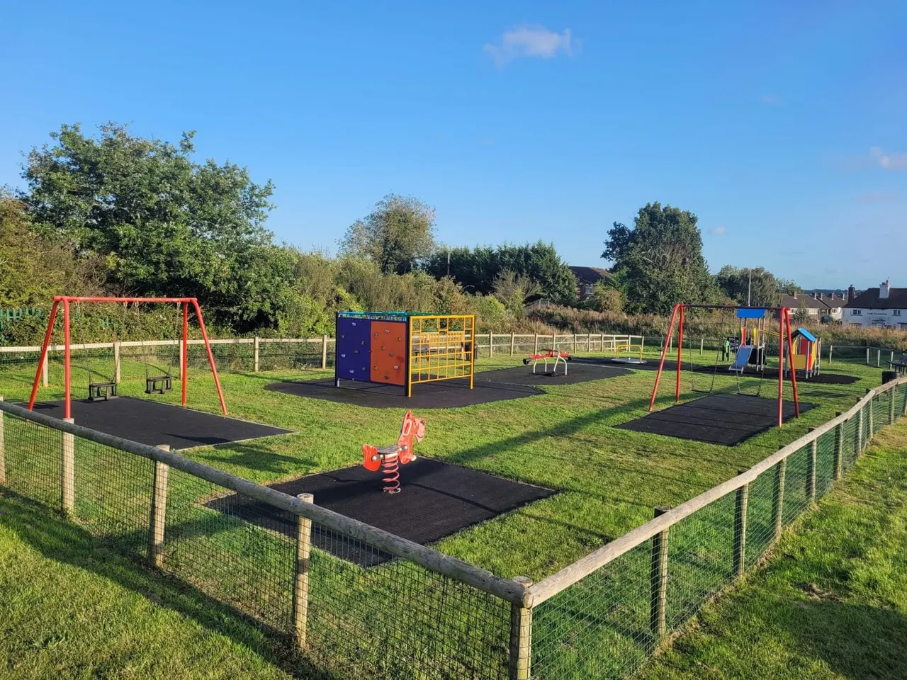 A children's play area with swings, climbing frames, slides and spring riders on dark safety surfaces, surrounded by grass and fencing.