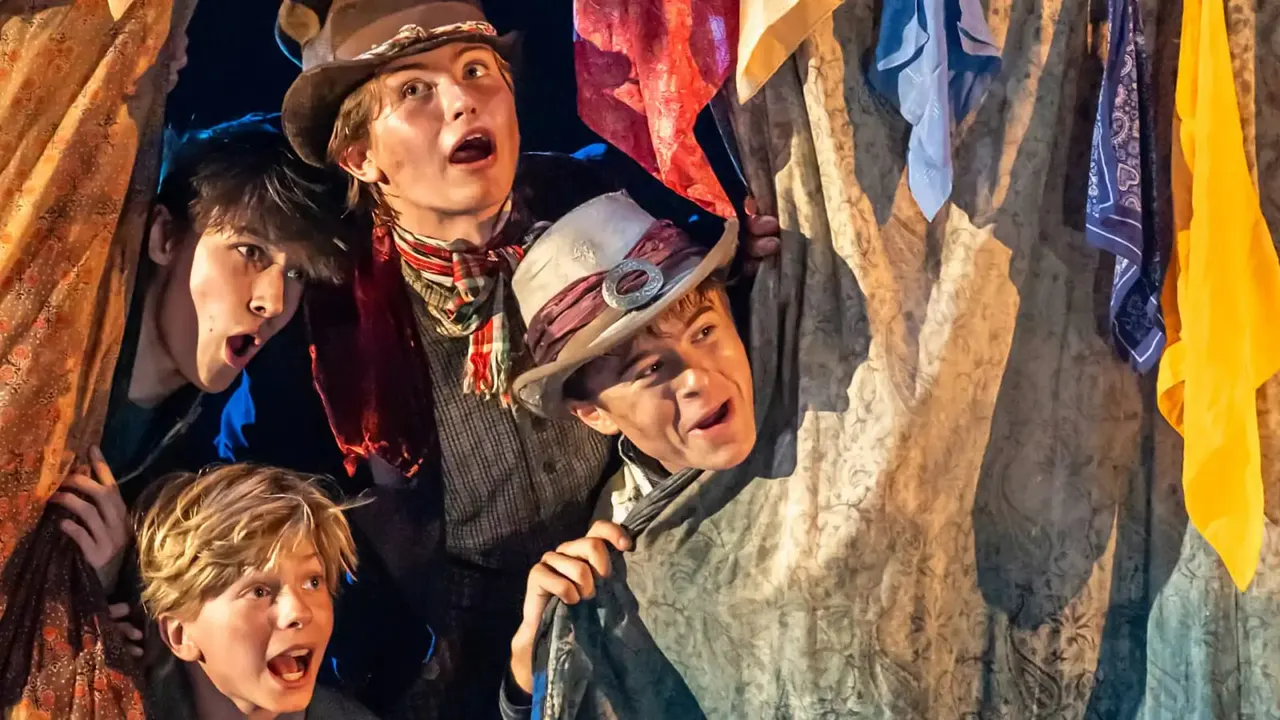 Four children in period costume stand among hanging fabrics and laundry in a theatrical scene.