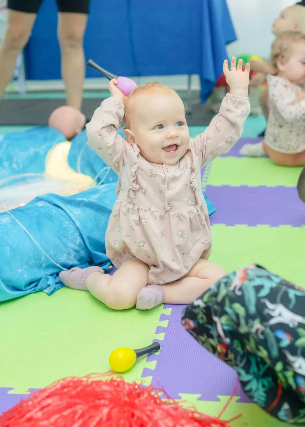 A smiling baby sits on a colourful padded mat during a class session, surrounded by toys and other infants.