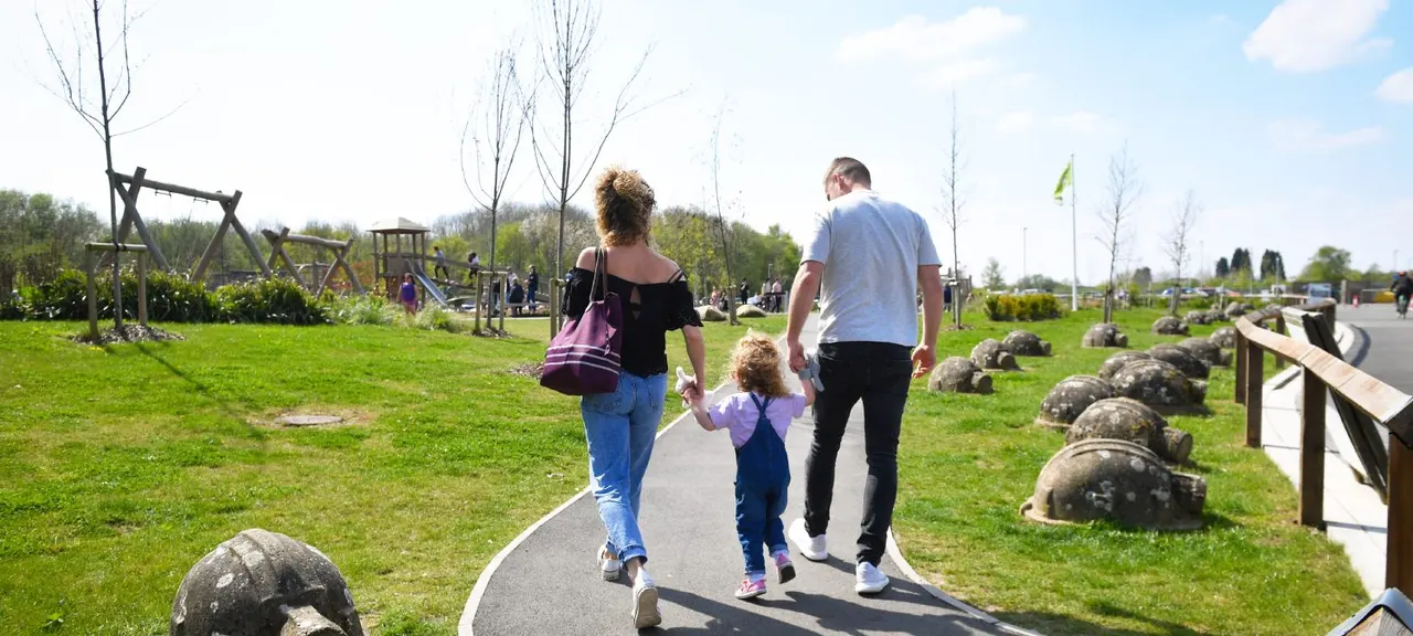 A woman, man and young child walk along a paved path through a sunny park with play equipment and landscaped grounds.