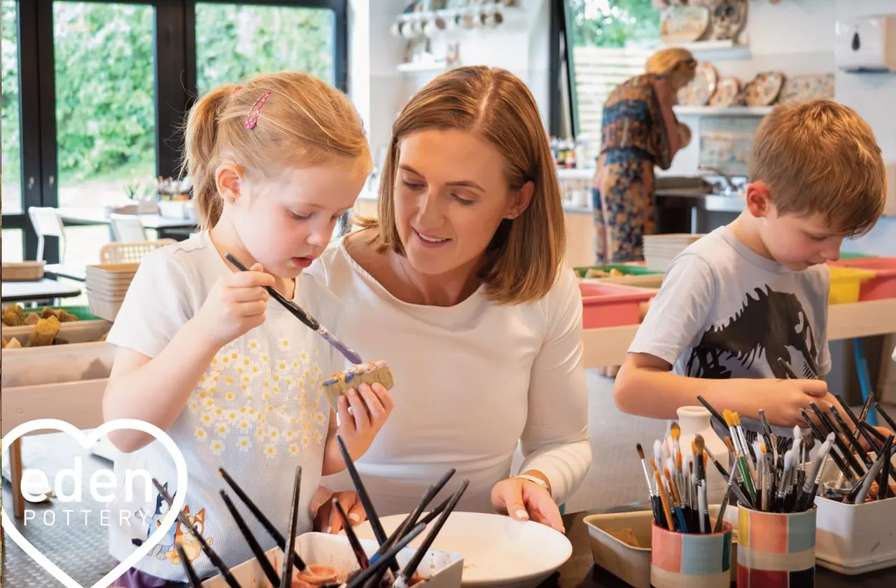 An adult and two young children painting pottery together at a bright studio table with brushes and paints.