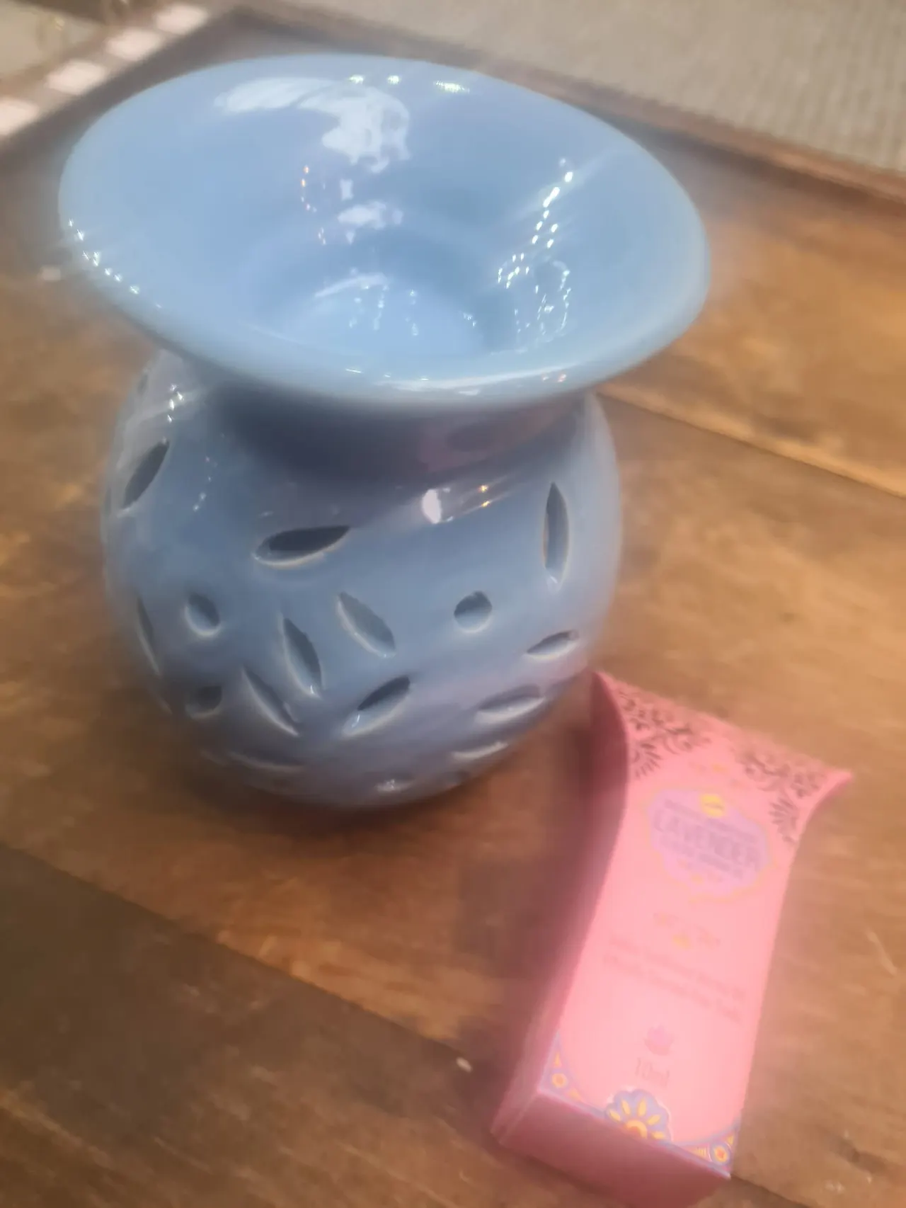 White ceramic oil burner with perforated body and bowl top, sitting on wooden surface next to pink box.