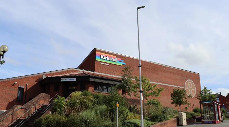 Exterior of a modern brick leisure centre building with signage, entrance stairs, and surrounding landscaping under a partly cloudy sky.