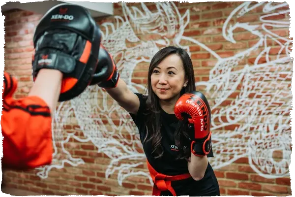 Woman in boxing gloves practising a punch against a brick wall with decorative artwork.