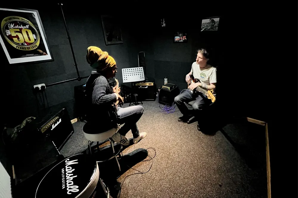 Two guitarists seated facing each other during a lesson in a dark music studio with Marshall amplifier and drum kit visible.