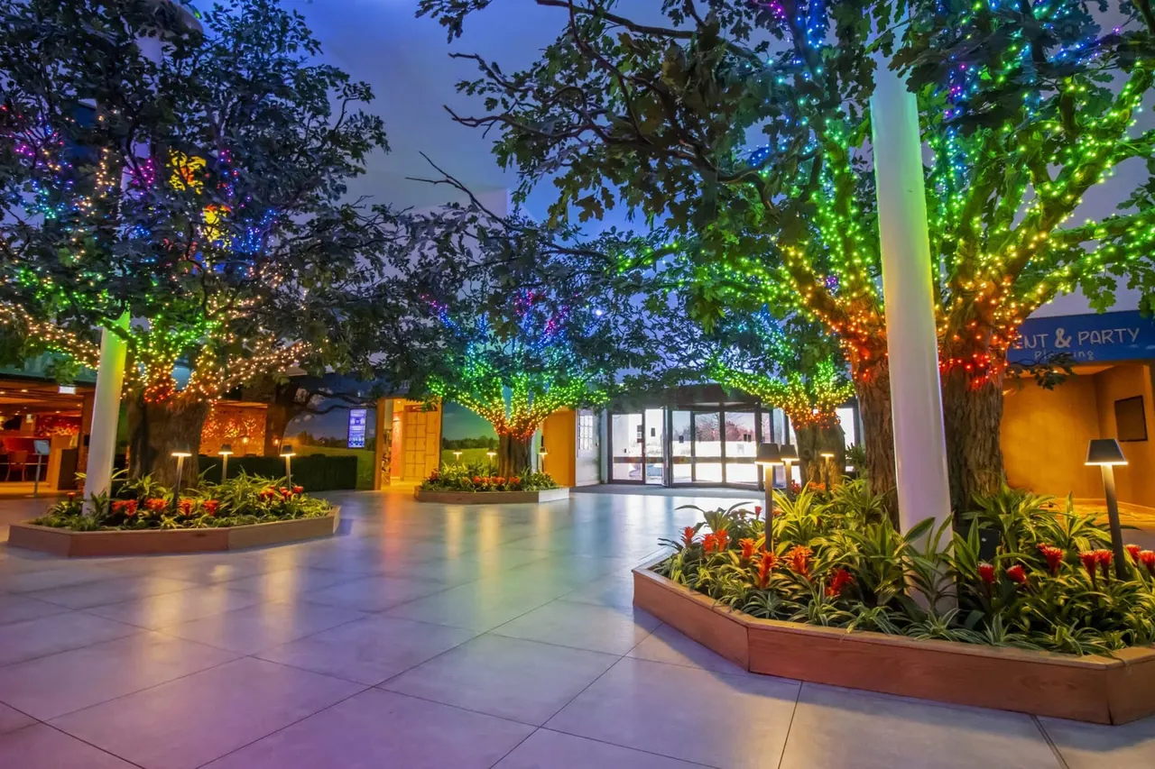 Outdoor entertainment venue courtyard at dusk with trees strung with multicoloured fairy lights, planted flower beds, and illuminated buildings surrounding a paved walkway.