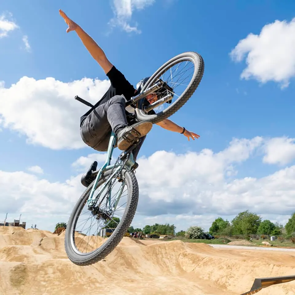 Cyclist performing a trick in the air above a dirt jump at a skate park against a blue sky.