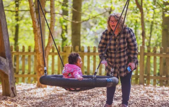 Adult pushing a young child on a tyre swing in a woodland playground area.