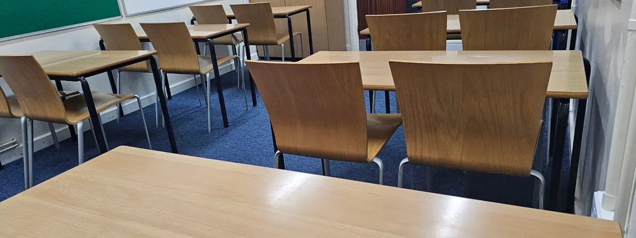 Empty classroom or community space with wooden tables and chairs arranged on blue carpet, with a green chalkboard visible on the wall.