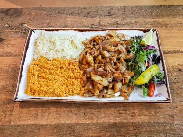 Plated meal with rice, yellow grain, grilled meat strips, and fresh vegetable garnish on wooden surface.