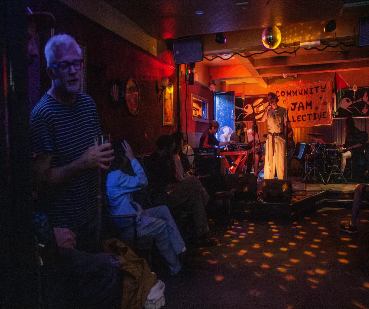 Band performing on stage at an indoor music venue with audience members watching in atmospheric red and blue lighting.