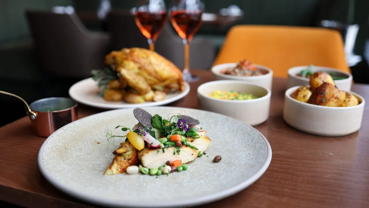 Fine dining table with plated fish dish garnished with vegetables and edible flowers, accompanied by side dishes, soups, roasted potatoes and two glasses of red wine.