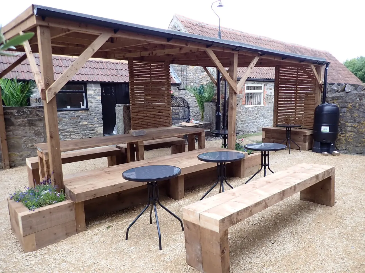 Outdoor wooden seating area with tables and benches under a timber pergola, adjacent to stone buildings.