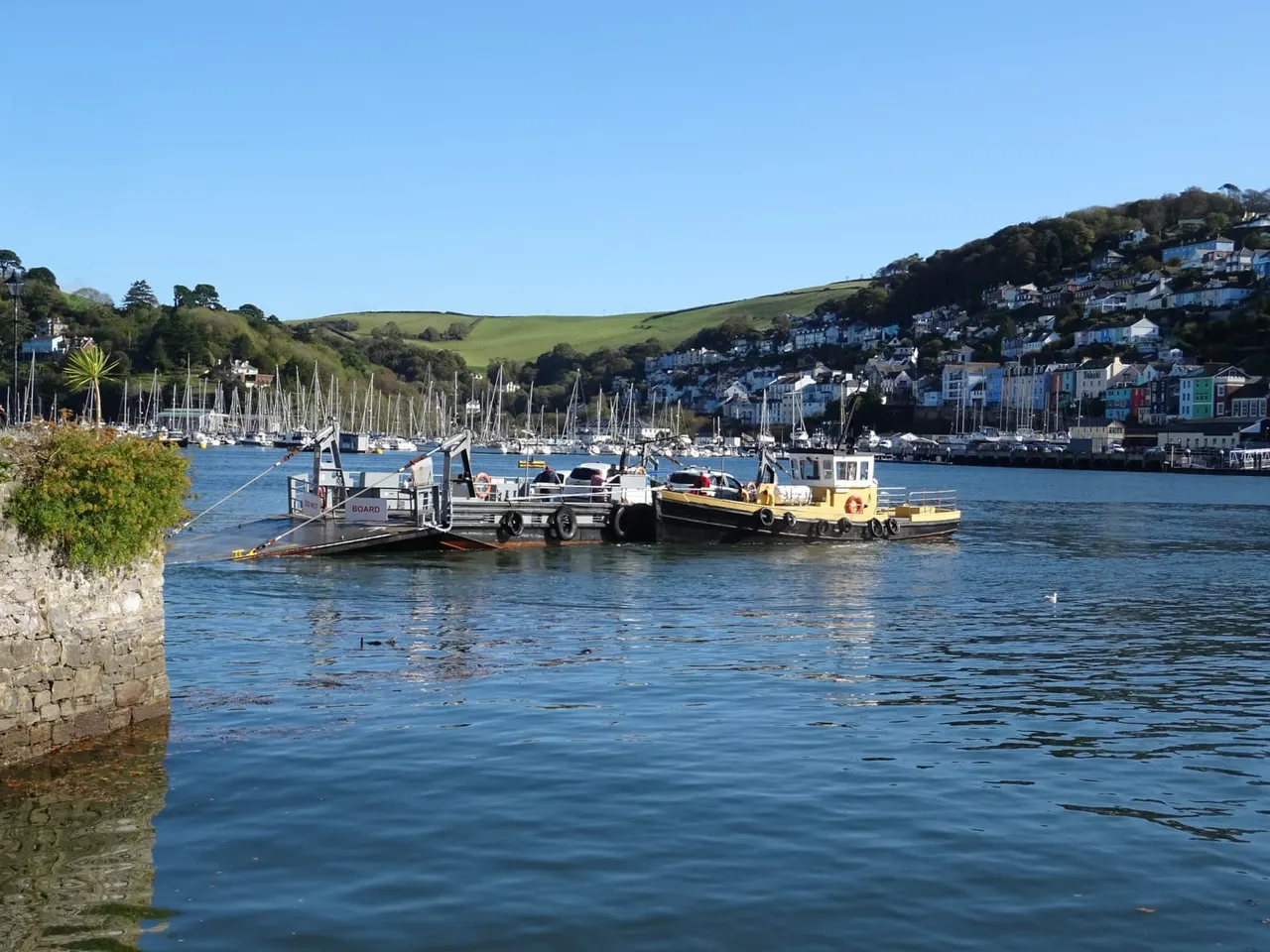 Car ferry loaded with vehicles moored on blue water with colourful waterfront houses and sailboats in background.