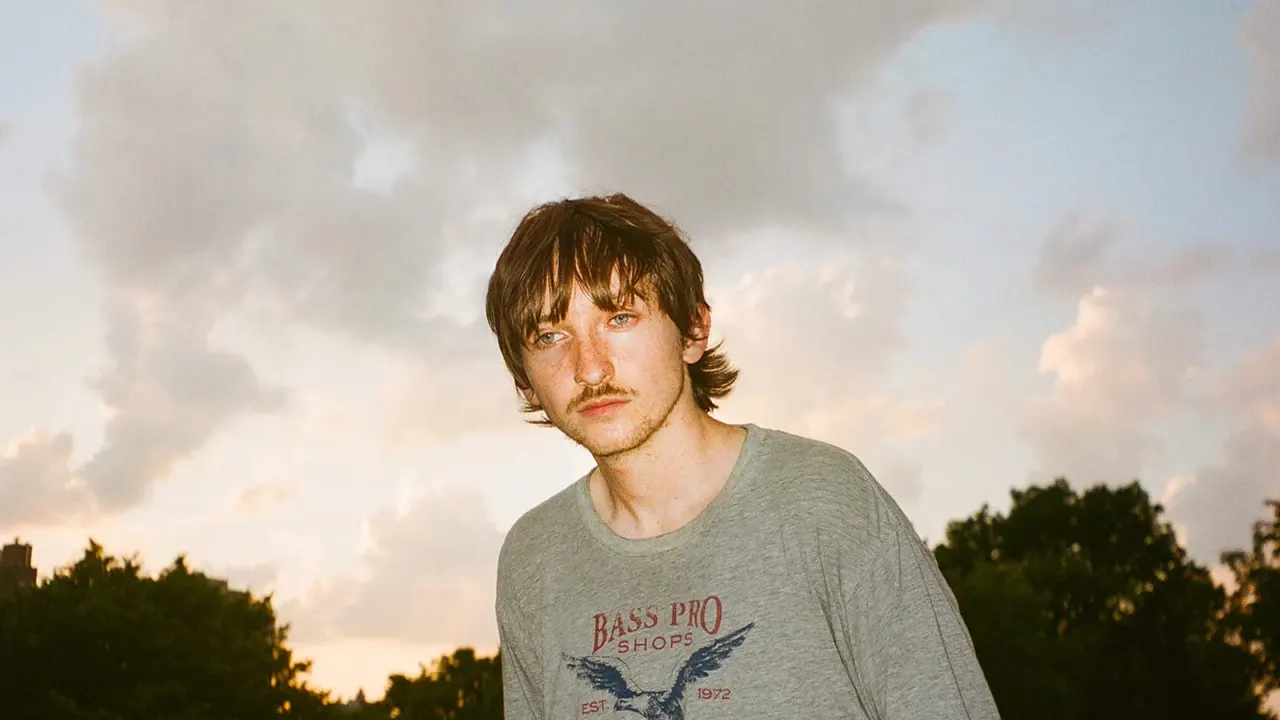 This is a photograph, not a designed graphic. Here is the alt text:

Man wearing a grey t-shirt stands outdoors against a cloudy sky with trees behind him.