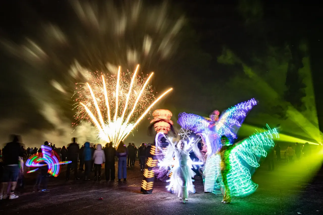 Crowd watching nighttime entertainment with fireworks exploding overhead and illuminated performers with glowing wings and props on ground.