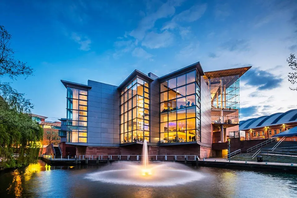 Modern concert hall with illuminated glass facade beside a canal with illuminated fountain, at dusk.