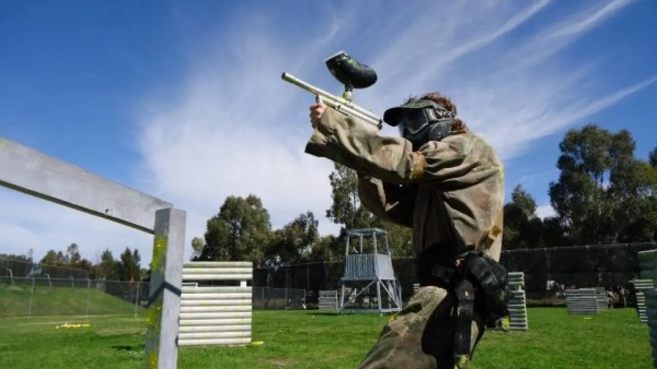 Player aiming paintball marker during outdoor game on grass field with obstacles.