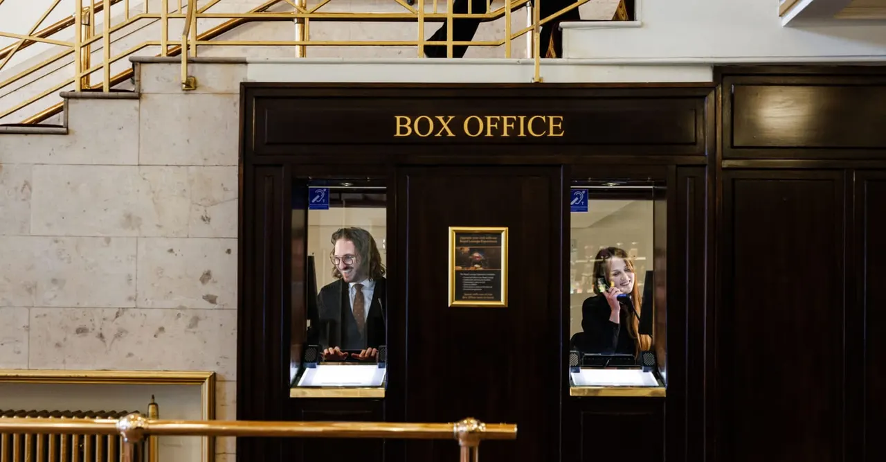 Box office counter with two staff members behind windows, gold and black signage, marble walls and brass railings.